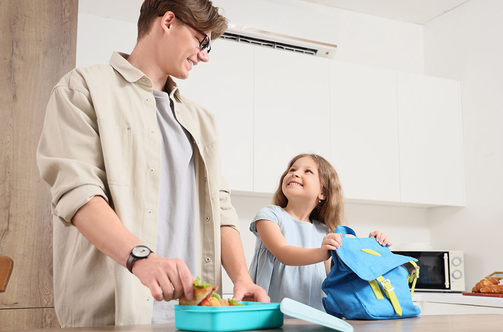 Az apa ebédet készít a lányának, aki mosolyogva tartja a hátizsákját.