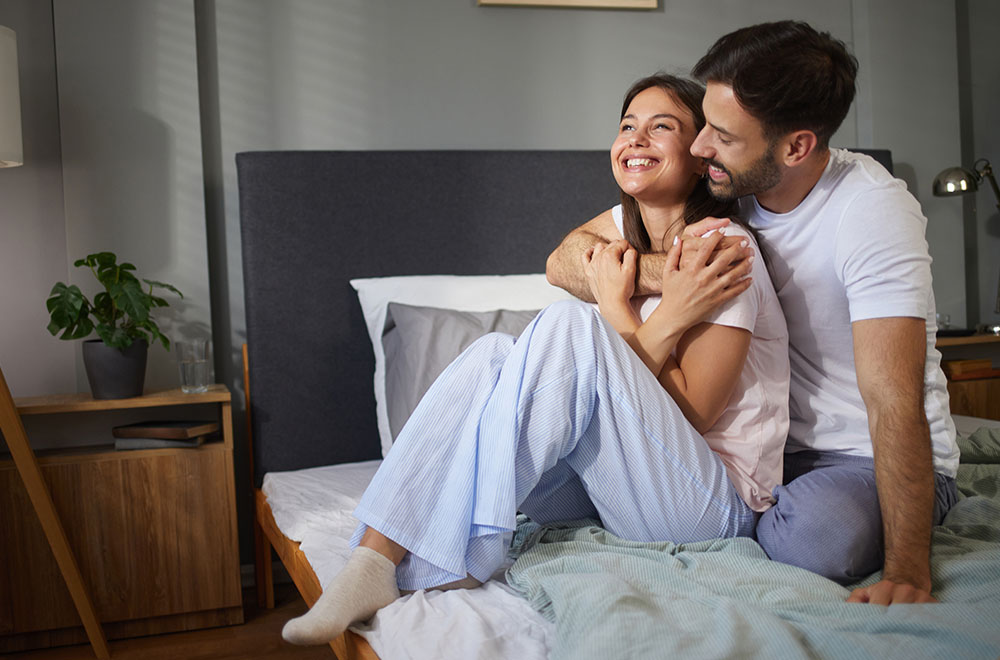 Egy pár, akik összebújva nevetnek az ágyban, osztozva egy kényelmes, boldog pillanaton.
