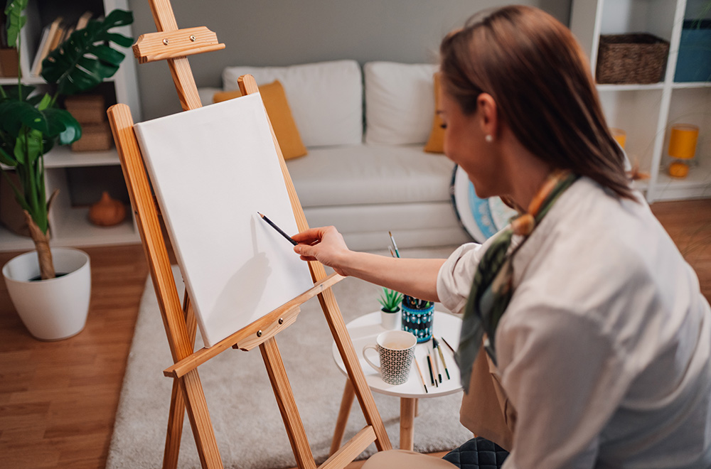 Egy nő ül egy üres vászon előtt a festőállványon, készen arra, hogy elkezdjen rajzolni, kezében ceruzát tartva.