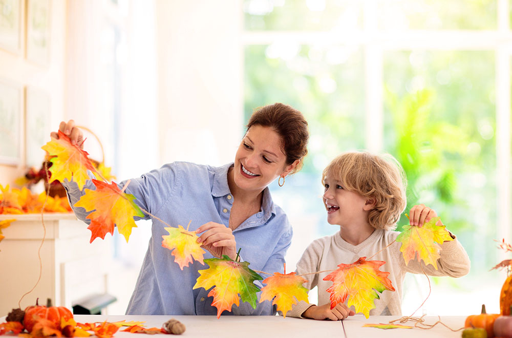 Anya és gyermeke színes levelekből dekorációt készítenek, mosolyogva egy világos, napos helyiségben.