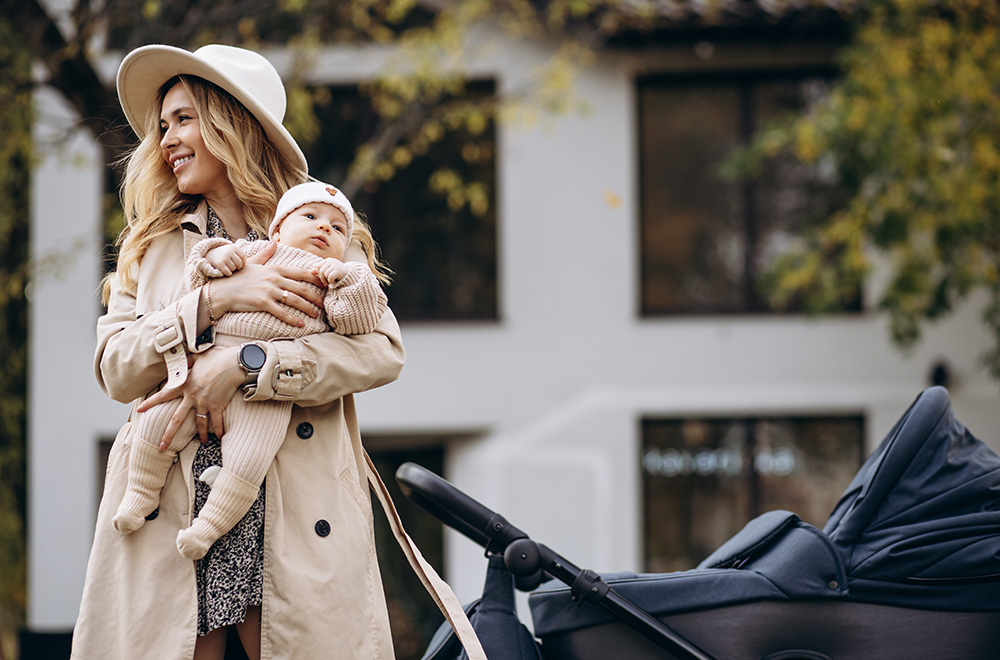 Egy nő karjában tart egy csecsemőt, miközben egy babakocsi mellett sétál az épület előtt.